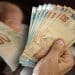 Mãos seguram notas de real representando a restituição de até R$ 1.000 paga via Pix pela Receita Federal