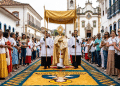 Procissão de Corpus Christi com sacerdote sob pálio dourado segurando o ostensório sobre tapete colorido em rua histórica