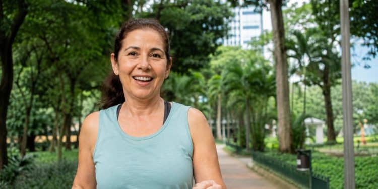 Mulher madura sorri ao caminhar em um parque arborizado, unindo exercício e contato com a natureza, dois hábitos do envelhecer bem.