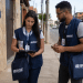 Dois profissionais fardados do IBGE realizam trabalho de campo em uma rua residencial, utilizando tablet e celular para coleta de informações.