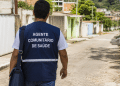 Agente Comunitário de Saúde caminha por rua residencial em imagem sobre vaga aberta na Prefeitura.