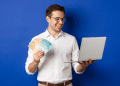 Homem sorrindo segura notas de real ao lado de notebook aberto em fundo azul.