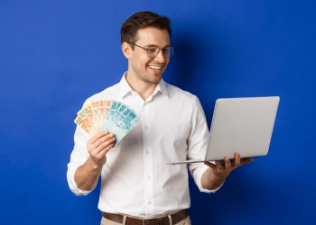 Homem sorrindo segura notas de real ao lado de notebook aberto em fundo azul.