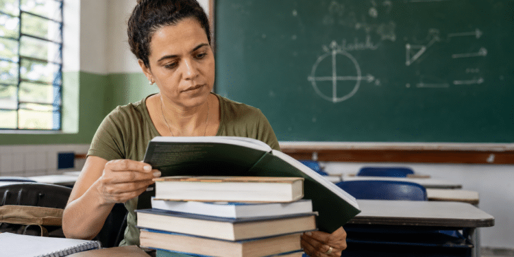 Professora folheia livro sobre pilha de obras em sala de aula com lousa ao fundo