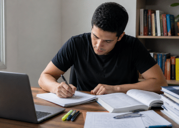 Pessoa estudando raciocínio lógico com notebook e livro aberto em mesa de estudos para concurso público.