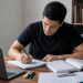 Pessoa estudando raciocínio lógico com notebook e livro aberto em mesa de estudos para concurso público.