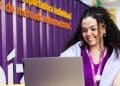 Jovem estagiária sorri trabalhando em notebook em escritório com parede roxa ao fundo