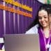 Jovem estagiária sorri trabalhando em notebook em escritório com parede roxa ao fundo