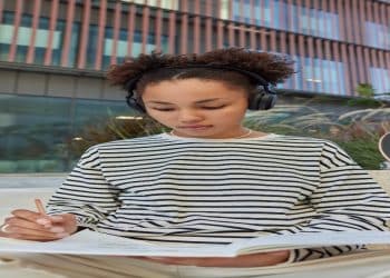 Jovem de fones de ouvido escreve concentrada em um caderno ao ar livre, representando a revisão de regras de português.