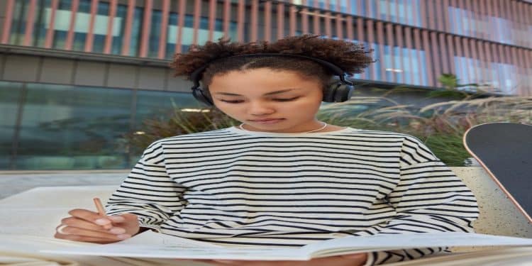 Jovem de fones de ouvido escreve concentrada em um caderno ao ar livre, representando a revisão de regras de português.