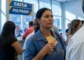 Mulher segura notas de dinheiro em uma fila dentro de uma agência da Caixa, sob um letreiro escrito PIS/Pasep, representando o pagamento do ressarcimento.