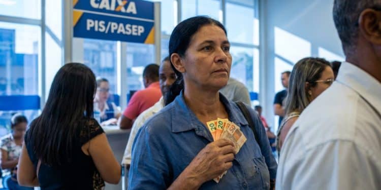 Mulher segura notas de dinheiro em uma fila dentro de uma agência da Caixa, sob um letreiro escrito PIS/Pasep, representando o pagamento do ressarcimento.