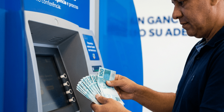 Homem conta cédulas de R$ 100 em frente a caixa eletrônico, representando o saque de até R$ 6.220 do FGTS por calamidade em 2026.
