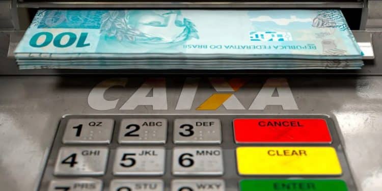 Cédulas de R$ 100 saindo de caixa eletrônico ilustram o Saque Calamidade do FGTS de até R$ 6.220 liberado pela Caixa