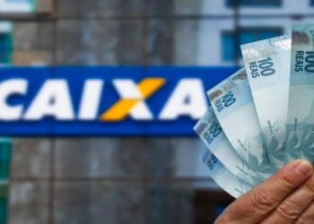 Mão segura cédulas de R$ 100 diante da fachada de uma agência da Caixa, banco que paga o saque calamidade do FGTS em junho.