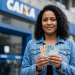 Mulher sorridente segura cédulas de real em frente a uma agência da Caixa, em referência ao saque de R$ 6.220 do FGTS.