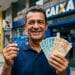 Homem sorri segurando cartão da Caixa e cédulas de R$ 100 e R$ 50 em frente a uma agência da Caixa, ilustrando o Saque Calamidade do FGTS de até R$ 6.220