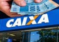 Mão segurando várias notas de R$ 100 sobre a fachada de uma agência da CAIXA, representando o pagamento do Pé-de-Meia.