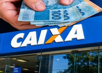 Mão segurando várias notas de R$ 100 sobre a fachada de uma agência da CAIXA, representando o pagamento do Pé-de-Meia.