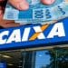 Mão segurando várias notas de R$ 100 sobre a fachada de uma agência da CAIXA, representando o pagamento do Pé-de-Meia.