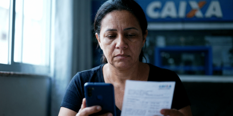 Mulher confere documento e celular em frente a uma agência da Caixa sobre o ressarcimento do PIS/Pasep