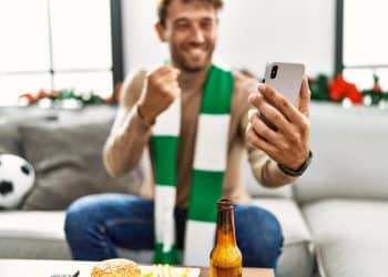 Torcedor com cachecol comemora ao acompanhar um jogo de futebol pelo celular em casa