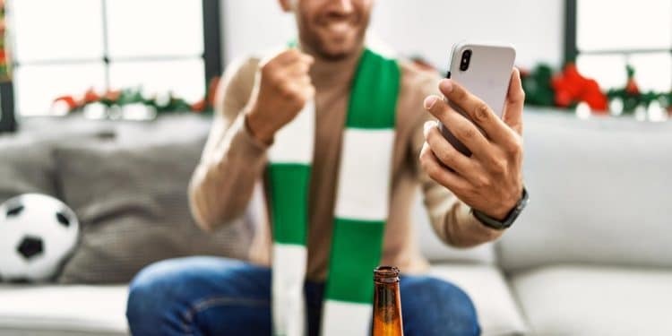 Torcedor com cachecol comemora ao acompanhar um jogo de futebol pelo celular em casa