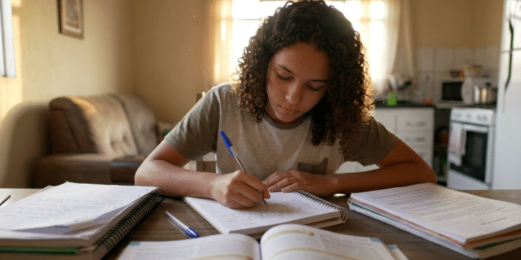 Jovem escreve em caderno cercada de livros e anotações sobre a mesa em casa