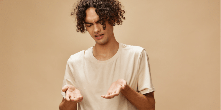 Jovem confuso observa as próprias mãos em fundo bege.