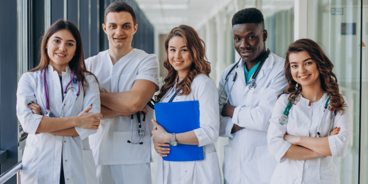 Equipe diversa de jovens profissionais da saúde sorrindo em corredor hospitalar.