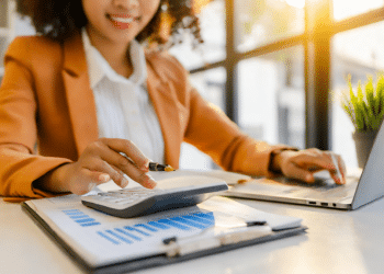 Jovem analisando finanças corporativas com calculadora e laptop em escritório iluminado.