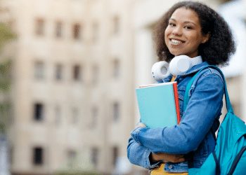 Estudante sorridente com livros e fones no campus universitário.