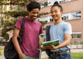 Dois jovens estudantes sorrindo, um negro e um indígena, lendo caderno ao ar livre.