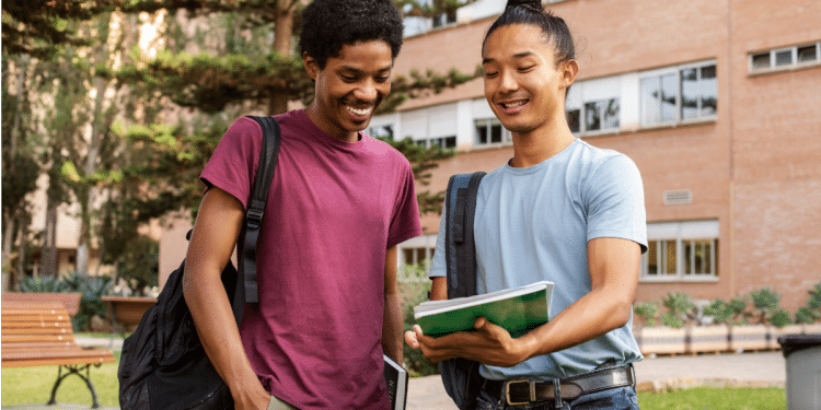 Dois jovens estudantes sorrindo, um negro e um indígena, lendo caderno ao ar livre.