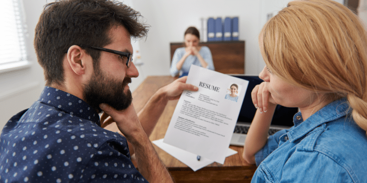 Homem mostra currículo impresso para mulher enquanto recrutadora observa ao fundo em escritório.