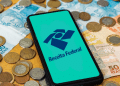 Smartphone com logo da Receita Federal sobre notas e moedas em fundo de madeira.
