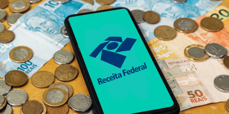 Smartphone com logo da Receita Federal sobre notas e moedas em fundo de madeira.