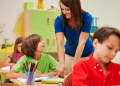 Professora orienta alunos em sala de aula colorida de ensino fundamental.
