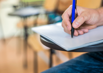 Estudante escrevendo em sala de aula com foco na mão.