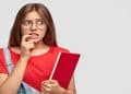 Jovem de óculos e expressão apreensiva, roendo a unha e segurando um livro vermelho, cena que representa a dúvida sobre o uso da vírgula na expressão "graças a Deus".