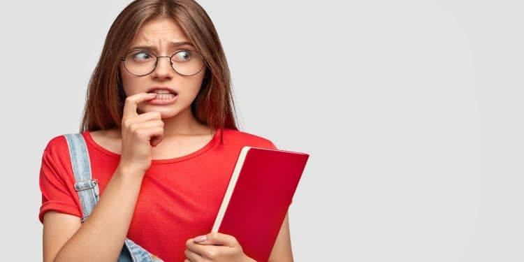 Jovem de óculos e expressão apreensiva, roendo a unha e segurando um livro vermelho, cena que representa a dúvida sobre o uso da vírgula na expressão "graças a Deus".