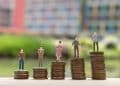 Figuras em miniatura sobre pilhas de moedas de tamanhos crescentes, representando diferenças salariais entre profissões