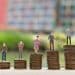 Figuras em miniatura sobre pilhas de moedas de tamanhos crescentes, representando diferenças salariais entre profissões
