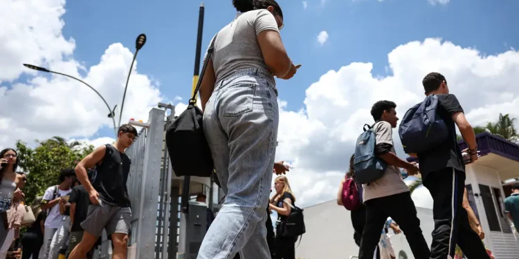 Candidatos com mochilas atravessam o portão de entrada do local de prova do Enem em dia ensolarado