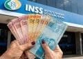 Idosa segurando cédulas de real em frente a uma agência do INSS, sobre o pagamento do 13º salário do INSS em 2026