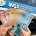 Idosa segurando cédulas de real em frente a uma agência do INSS, sobre o pagamento do 13º salário do INSS em 2026