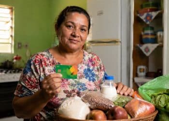 Mulher segura um cartão alimentação ao lado de uma cesta com arroz, feijão, frutas, legumes e leite, ilustrando os programas estaduais que pagam de R$ 100 a R$ 300 por mês
