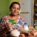 Mulher segura um cartão alimentação ao lado de uma cesta com arroz, feijão, frutas, legumes e leite, ilustrando os programas estaduais que pagam de R$ 100 a R$ 300 por mês