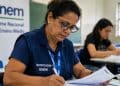 Mulher com colete de certificador do Enem preenche um relatório em uma sala, ao lado de um banner do Exame Nacional do Ensino Médio, representando a função exercida na aplicação da prova
