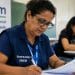 Mulher com colete de certificador do Enem preenche um relatório em uma sala, ao lado de um banner do Exame Nacional do Ensino Médio, representando a função exercida na aplicação da prova